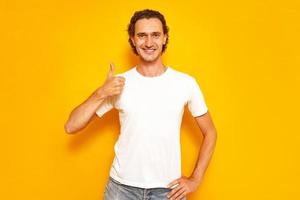 young handsome man in casual T-shirt, standing on an isolated yellow background, makes happy thumbs up gesture with his hand. approving expression of face looking into camera shows success. copy space photo