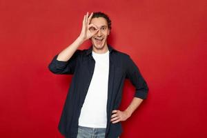 agitated man brings his hand to his eye showing ok sign. dressed in casual clothes. isolated on red background with space for advertising text. concept - people, body language, luck, victory, planning photo