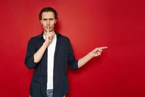 serious man puts his finger to his lips and points towards empty space for advertising. studio portrait. dressed in casual clothes. isolated on red background. concept - taboo, secret sale, mystery photo