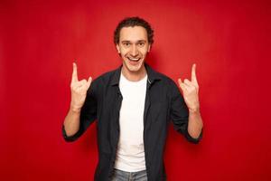 Happy overjoyed beautiful male with curly hair, shows cool sign love, says Yeah, that wonderful Pretty young man gestures indoor against red background copy space. People and body language concept photo