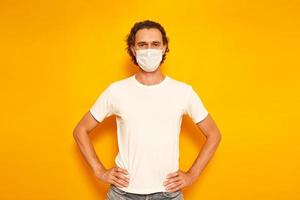 curly-haired young man in casual clothes wearing protective medical mask stands with his hands on his belt isolated yellow background with space for text. concept people, protection from virus, health photo