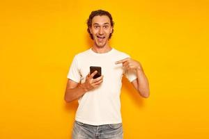an excited happy man in casual clothes with phone in his hand points his finger at smartphone and we are happy to receive message about discounts, promotions and sales. isolated on yellow background. photo