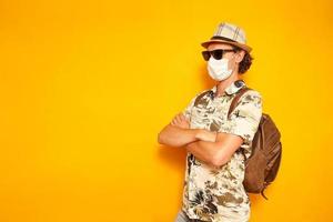 male tourist traveler in medical protective mask, sunglasses, with backpack on vacation crossed his arms on his chest looks at an empty area. isolated on yellow background with space for text photo