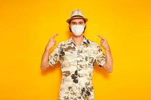 male tourist traveler points with his index fingers at a medical mask on his face. isolated on a yellow background with space for text. tconcept of people, vacation, protection from virus covid-19 photo