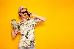 satisfecho viajero turista masculino feliz con bebida tropical en la mano está hablando por teléfono y disfrutando de vacaciones en el extranjero. fondo amarillo aislado con espacio para texto. concepto recreación comunicación foto