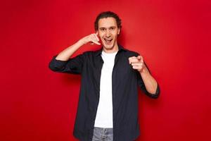 call me hand sign. portrait of young smiling man pointing index finger in front of him depicts telephone receiver in his hand. isolated on red studio background. concept - love, romance, communication photo