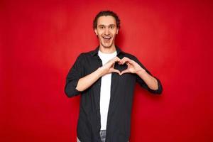 cheerful excited man shows heart sign with hands in front of his chest, confesses his love with his mouth open. isolated on red background copy space. concept - romance, relationships, valentine's day photo