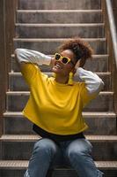 Happy black hipster woman in trendy wear and sunglasses sitting on steps photo