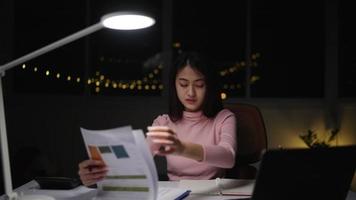 carreira freelance de mulher asiática vestindo roupas cor de rosa fazendo papelada há um laptop em cima da mesa. e lanternas acesas, deliberadamente ficando em casa durante a noite galesa video