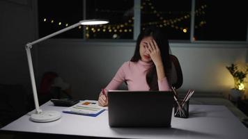 slow-motion, aziatische freelance vrouw draagt roze kleren terwijl ze overwerkt is. slaperig en probeerde 's nachts met laptop op bureau en lamp thuis te schrijven. concept mensen freelance werken in de loop van de tijd. video