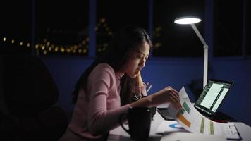 mujer independiente asiática usa ropa rosa comiendo fideos preparados mientras trabaja demasiado. escriba con la computadora portátil en el escritorio y la lámpara en casa por la noche. concepto de trabajo independiente de las personas con el tiempo.