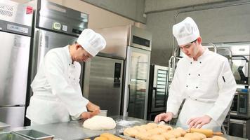 Two professional Asian male chefs in white cook uniforms and aprons are kneading pastry dough and eggs, preparing bread and fresh bakery food, baking in oven at stainless steel kitchen of restaurant.