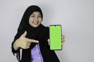 Young Asian Islam woman wearing headscarf is showing on green screen of phone with happy in blurry background. photo