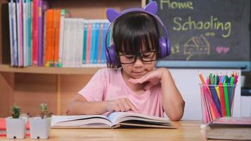 menina bonitinha com fones de ouvido ouvindo audiolivros e olhando livros de aprendizagem de inglês em cima da mesa. aprendendo inglês e educação moderna video
