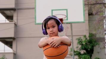 una linda niñita con auriculares posa con baloncesto en una cancha de baloncesto al aire libre.