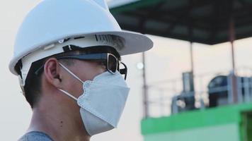 joven ingeniero asiático con casco y máscara mira y sonríe a la cámara en el fondo de la presa. un joven ingeniero trabaja en una presa durante el brote de coronavirus. video