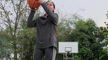 atleta feminina asiática usando poses de fones de ouvido com basquete na quadra de basquete ao ar livre.