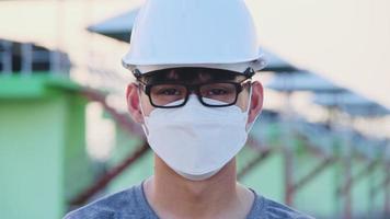 jovem engenheiro asiático usando um capacete e máscara olha e sorri para a câmera no fundo da barragem. um jovem engenheiro trabalha em uma barragem durante o surto de coronavírus.