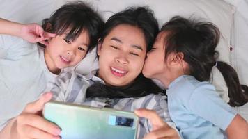felice famiglia asiatica godendo con lo smartphone a casa accogliente. madre sorridente e figlie carine usando il telefono, fai un selfie o un video clip da ricordare a letto a casa. buon tempo a casa