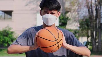 joven asiático con una máscara facial sosteniendo una pelota de baloncesto en las manos en una cancha al aire libre durante el brote de covid-19.