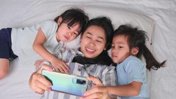 feliz família asiática desfrutando com smartphone em casa aconchegante. mãe sorridente e filhas fofas usando telefone, tire uma selfie ou videoclipe para lembrar na cama em casa. bom tempo em casa