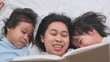 adorável família asiática feliz em casa aconchegante. muito jovem mãe lendo um livro para suas filhas em uma cama em casa. bom tempo em casa. video