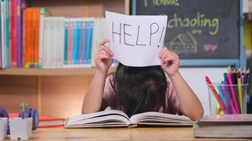 Aziatisch klein meisje dat zich verdrietig en moe voelt terwijl ze aan tafel zit met veel boeken en een papier vasthoudt met woordhulp. leer moeilijkheden. thuisonderwijs. video