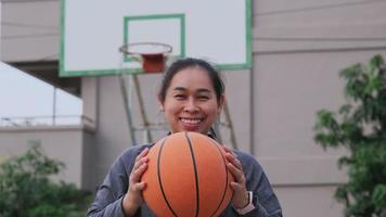 aziatische vrouw die basketbal houdt die camera bekijkt bij openluchtbasketbalspeelplaats. video