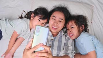 glad asiatisk familj njuter med smartphone i mysiga hem. leende mamma och söta döttrar använder telefonen, ta en selfie eller videoklipp för att minnas i sängen hemma. bra tid hemma video