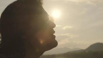 Handsome curly haired Asian man playing guitar and singing by the lake at sunset on vacation. video