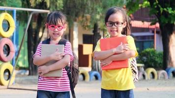 twee schattige schoolmeisjes die zomerkleren dragen met rugzakken die een boek vasthouden en glimlachen op school. terug naar school concept video