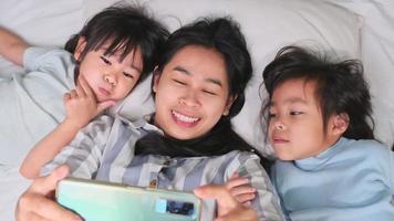 Happy Asian family enjoying with smartphone at cozy home. Smiling mother and cute daughters using phone, Take a selfie or video clip to remember in bed at home. Good time at home