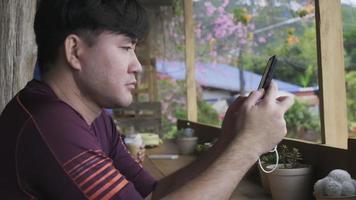 asiatischer mann, der in einem café sitzt und eine nachricht auf einem handy liest. Schöner Mann mit Smartphone im Café. video