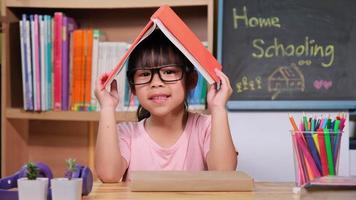 linda niña sosteniendo un libro sobre su cabeza como un techo, sonriendo y mirando a la cámara. adorable niño leyendo un libro para la educación en el hogar. video