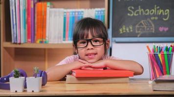 una niña linda sonríe y pone su barbilla sobre su mano en una pila de libros en casa. adorable niño leyendo un libro para la educación en el hogar. video