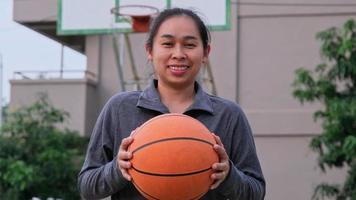atleta asiática con auriculares posa con baloncesto en la cancha de baloncesto al aire libre.