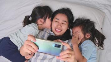 gelukkige aziatische familie genieten met smartphone in gezellig huis. lachende moeder en schattige dochters die telefoon gebruiken, maak een selfie of videoclip om thuis in bed te onthouden. lekker thuis video