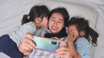 famille asiatique heureuse appréciant avec smartphone dans une maison confortable. mère souriante et filles mignonnes utilisant le téléphone, prenez un selfie ou un clip vidéo à retenir au lit à la maison. bon moment à la maison video