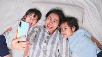 famille asiatique heureuse appréciant avec smartphone dans une maison confortable. mère souriante et filles mignonnes utilisant le téléphone, prenez un selfie ou un clip vidéo à retenir au lit à la maison. bon moment à la maison video