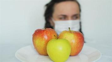 mujer con mascarilla médica rociando manzanas con productos químicos y poniendo un plato con la palabra gmo. enfoque selectivo en la mujer y luego en las manzanas. concepto de fruta genéticamente modificada video