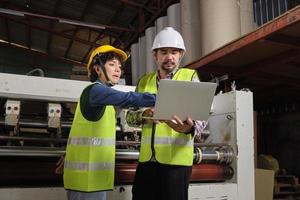 Safety uniform workers and industrial engineers with hardhat use laptop computer to check and control machines. Two professionals work in paper manufacturing factory, maintain production equipment. photo