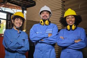 retrato de tres ingenieros industriales multirraciales del equipo de trabajadores con uniforme de seguridad y casco protector miran la cámara, cruzan el brazo y sonríen felizmente en el almacén de la fábrica de cartón. foto