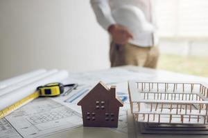 Engineer is holding a helmet standing in his office and has a blueprint and model on his desk. photo