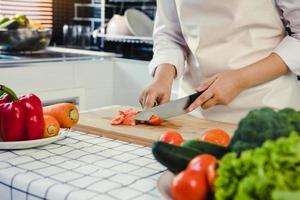 Woman using knife and hands cutting tomato on wooden board in kitchen room.