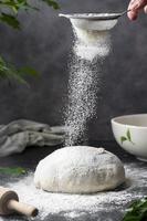 Cooking sourdough homemade bread. Sieving flour over raw bread dough on table, dark background, selective focus. photo