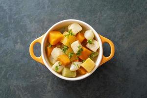 Root vegetables casserole in orange bowl, with fresh herbs. Vegan food. Dark background, top view. photo