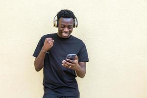 alegre joven negro apretando el puño y sonriendo mientras usa el teléfono inteligente en la calle foto