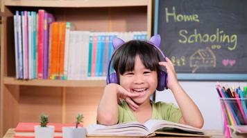 menina bonitinha com fones de ouvido ouvindo audiolivros e olhando livros de aprendizagem de inglês em cima da mesa. aprendendo inglês e educação moderna video