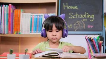 Cute little girl with headphones listening to audiobooks and looking at English learning books on the table. Learning English and modern education video