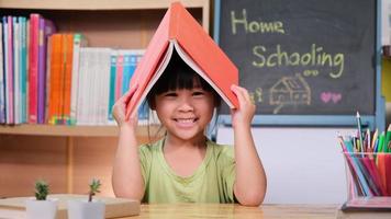 jolie petite fille tenant un livre au-dessus de sa tête comme un toit, souriant et regardant la caméra. adorable enfant lisant un livre pour l'école à la maison. video